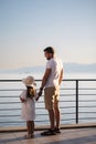 Back view of daughter with her father stand on heigh balcony in front of sea sunset. Dad with his little girl looking on
