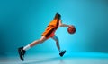 Back view dynamic image of young girl, basketball player dribbling ball against blue studio background in neon light