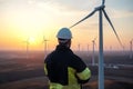 Back view of engineer wearing safety helmet and reflective vest standing on wind turbine field at sunset. Generative AI