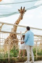 Back View Of Father And Daughter In Straw Hat Standing Near Fence And Giraffe In Zoo.