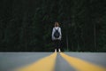 Back view on a female hiker walking down the asphalt road in the woodland. Tourist woman with a backpack standing in the middle of