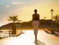 Back view of female silhouette going on evening sunset background of resort hotel. Woman walking by tropic street way with palms