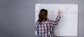 Back view of female teacher standing at a whiteboard, writing on the board, explaining grammar rules to students