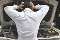 Back view of frustrated stressed young mechanic man in white uniform touching his head with hands against car in open hood at the Royalty Free Stock Photo