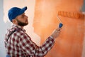 Back view of handsome painter in blue dungarees painting a wall with paint roller in orange color. Royalty Free Stock Photo