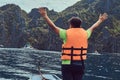 Back view of happy guy in a life jacket stands on a boat on the background of a beautiful landscape of rocky bay.