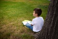 Back view of happy schoolboy resting after school , sitting on the green grass of the city park, leaning against a tree, doing his Royalty Free Stock Photo