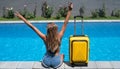 Back view of happy woman with yellow luggage sitting by the swimming pool