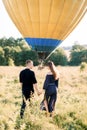 Back view of happy young woman and man in summer field, ready to make balloon tour, standing in front of air balloon Royalty Free Stock Photo