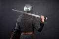 Back view of a knight in chain mail and a helmet with a sword on his shoulder on a dark background. Royalty Free Stock Photo