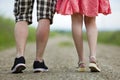 Back view of legs of young slim woman in red dress and man in shorts walking together by ground road on sunny summer day on Royalty Free Stock Photo