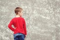 Back view of little boy looking at wall. Rear view on gray concrete wall. Mockup. Cool boy wearing red shirt and jeans. Back to