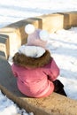 Back view of little girl in warm winter clothes sitting on low concrete circle in park