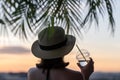 Back view of lonely beautiful girl with glass of champagn in a straw hat against the background of the sea in branches of palm