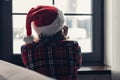 Back view of lonely sad young woman in a red santa claus christmas hat sitting and looking at window. Royalty Free Stock Photo