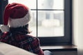 Back view of lonely sad young woman in a red santa claus christmas hat sitting and looking at window. Royalty Free Stock Photo