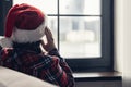 Back view of lonely sad young woman in a red santa claus christmas hat sitting and looking at window. Royalty Free Stock Photo