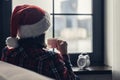 Back view of lonely sad young woman in a red santa claus christmas hat sitting near window with alarm clock. Concept. Royalty Free Stock Photo