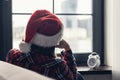 Back view of lonely sad young woman in a red santa claus christmas hat sitting near window and holding alarm clock. Royalty Free Stock Photo