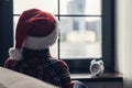 Back view of lonely sad young woman in a red santa claus christmas hat sitting near window and holding alarm clock. Royalty Free Stock Photo