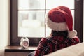 Back view of lonely sad young woman in a red santa claus christmas hat sitting near window and holding alarm clock. Royalty Free Stock Photo