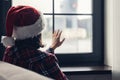 Back view of lonely woman in a red santa claus christmas hat sitting, looking and touching window. Royalty Free Stock Photo