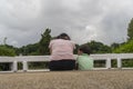 Back view of loving Asian mother hug her kids sitting on bridge, caring black mom embrace child, relaxing looking to black cloud, Royalty Free Stock Photo