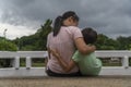 Back view of loving Asian mother hug her kids sitting on bridge, caring black mom embrace child, relaxing looking to black cloud, Royalty Free Stock Photo