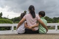 Back view of loving Asian mother hug her kids sitting on bridge, caring black mom embrace child, relaxing looking to black cloud, Royalty Free Stock Photo