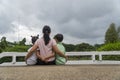 Back view of loving Asian mother hug her kids sitting on bridge, caring black mom embrace child, relaxing looking to black cloud, Royalty Free Stock Photo