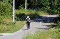 Back view of male person in white shirt with backpack and baseball cap riding a bicycle on local paved road surrounded with grass