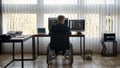 Back view of male web developer in a wheelchair writing program code on multiple computer screens while sitting at his Royalty Free Stock Photo