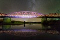 Back view of man with flashlight standing on river bank under illuminated metal bridge under dark starry sky reflected in water.