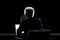 Back view of a man with gray hair working from home late at night talking on his phone while using his laptop computer Royalty Free Stock Photo