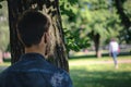 Back view of man stalking woman while hiding behind tree in public park with blurry woman in background Royalty Free Stock Photo