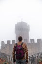 Back view of man wearing leather jacket and backpack. Fortification, old castle on background. Journey. Mystical atmosphere, fog,