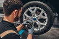 Back view of mechanic is repairing car at service station. Close up view of repairman is removing wheel by electric wrench, tyre