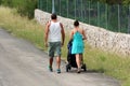 Back view of modern young couple pushing stroller uphill on side of paved road next to uncut grass and traditional stone wall