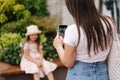 Back view of mom takes photos of her daughter on her phone. Adorable little girl sitting on bench. Focus mother& x27;s