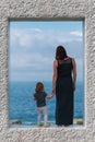 Back view on mother with daughter who stay on marble frame and looking at the sea