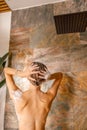 Back view of naked young woman washing her hair using organic natural shampoo while taking shower in marble bathroom