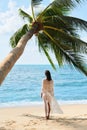 Back view of pretty young woman relax and enjoy sea standing under palm tree on tropical beach