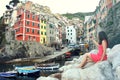 Back view of red dressed girl sitting on the stones like a mermaid taking photo with mirrorless camera of Italian Riviera landscap