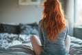 Back view of a redhaired woman sitting on the edge of her bed.