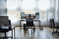 Back view of relaxed young male trader in a wheelchair keeping hands behind head and looking at three computer monitors