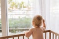 Back view of sad lonely baby standing in bed looking through window waiting for his mom Royalty Free Stock Photo