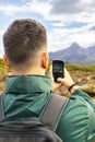 Back view shot of a young caucasian man wearing a green jacket and a backpack taking a photo with his smarphone of the Scottish Royalty Free Stock Photo