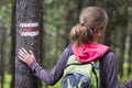 Back view of slim tourist hiker girl with stick and backpack holding hand on pine tree trunk with way sign in lit by sun mountain Royalty Free Stock Photo