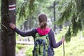 Back view of slim tourist hiker girl with stick and backpack holding hand on pine tree trunk with way sign in lit by sun mountain Royalty Free Stock Photo