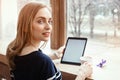 Back view smiling beautiful young woman read magazine with digital tablet near big window in a cafe or home. Attractive Royalty Free Stock Photo